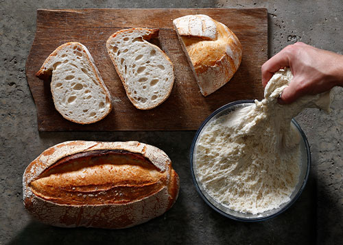 Ein Brot, ein angeschnittenes Brot und eine Schale mit LIEVITO MADRE PURO ATTIVO Sauerteig sind angerichtet platziert 